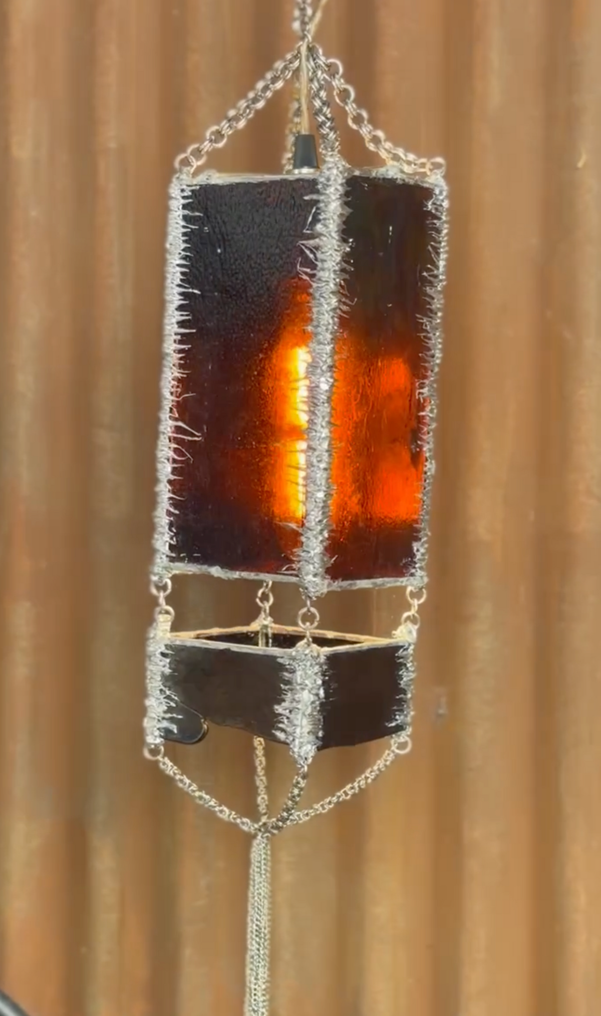 A hanging lantern-style pendant lamp with a rectangular amber glass panel, silver chain mail framing, and a smaller matching panel below, shown against a warm brown wood-textured background.