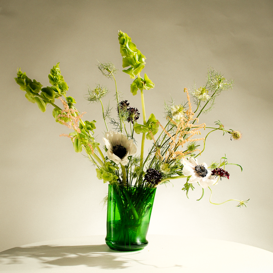 A green glass vase filled with a mixed floral arrangement featuring green leaves, white anemone-like flowers, and delicate dried stems, set against a plain light beige studio background.