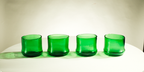 Four green translucent glass tumblers arranged in a row on a white surface against a light beige background.