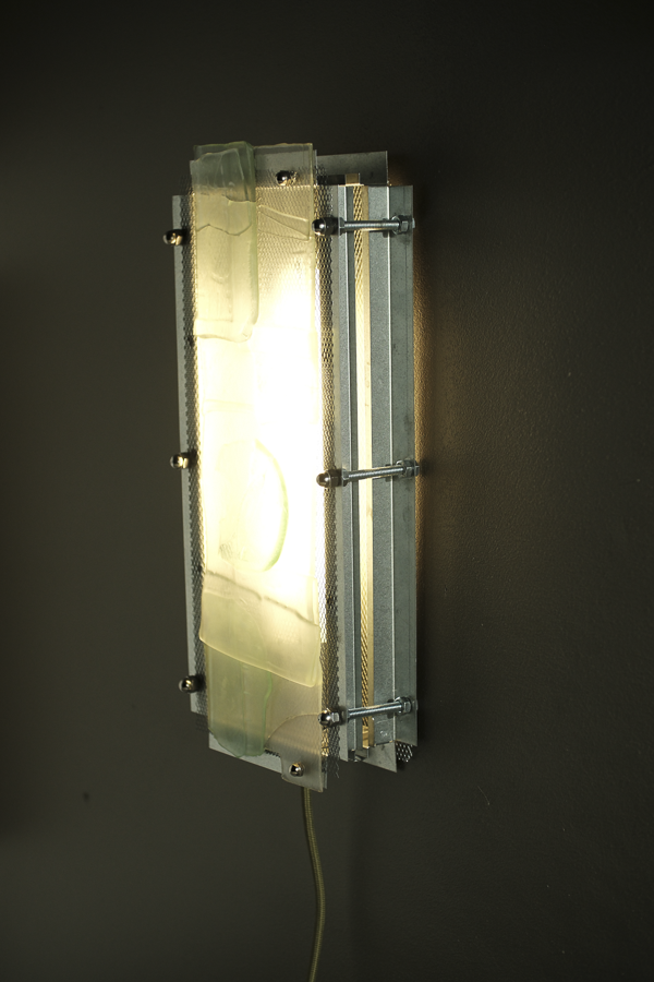 A modern industrial wall sconce with a glowing rectangular glass shade, mounted on a dark wall. The fixture features a brushed silver metal frame with visible screws and horizontal mounting brackets.