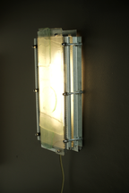 A modern industrial wall sconce with a glowing rectangular glass shade, mounted on a dark wall. The fixture features a brushed silver metal frame with visible screws and horizontal mounting brackets.