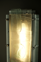 A rectangular translucent glass panel mounted on a metal frame with visible screws and mesh backing, glowing with warm light against a dark background.