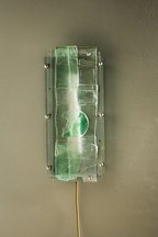 A rectangular wall-mounted light fixture with a translucent green glass shade, a white diffuser, and a thin yellow cord extending downward, shown against a plain taupe wall.