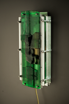A rectangular wall-mounted light fixture with a translucent green glass panel, silver metal frame, and visible screws, shown against a dark gray wall.