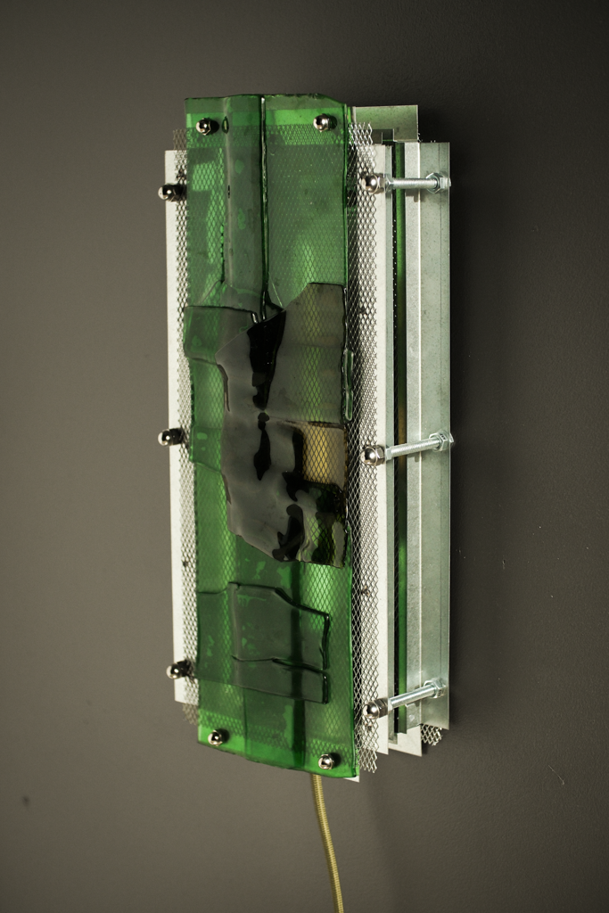 A rectangular wall-mounted light fixture with a translucent green glass panel, silver metal frame, and visible screws, shown against a dark gray wall.