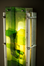 A close-up of a modern wall light fixture featuring translucent green and yellow glass panels mounted on a brushed silver metal frame with visible screws and mesh details.