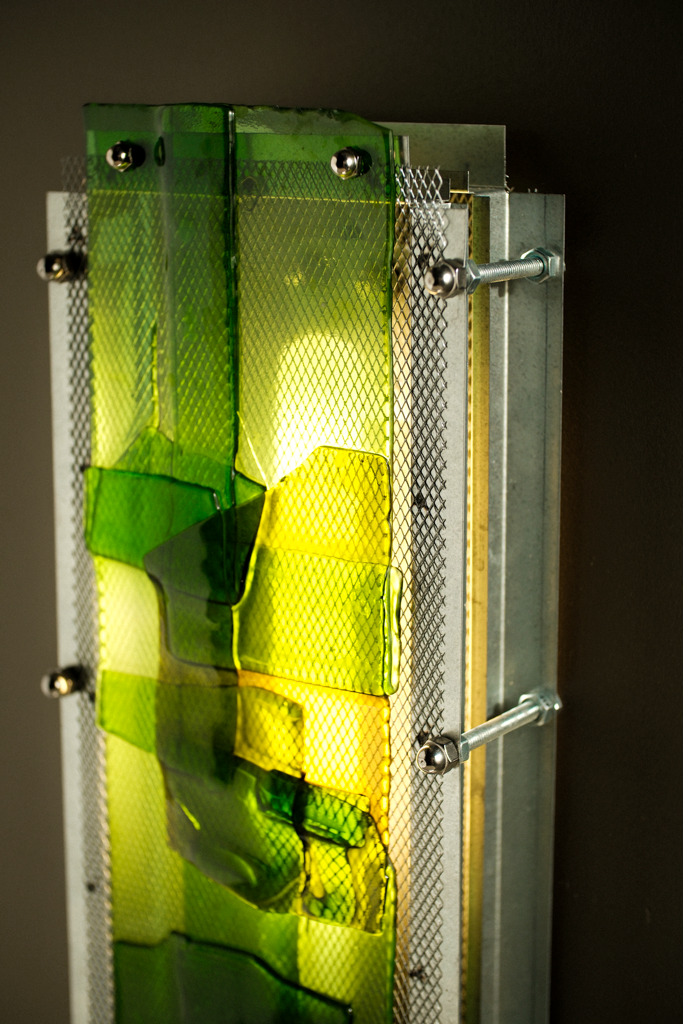 A close-up of a modern wall light fixture featuring translucent green and yellow glass panels mounted on a brushed silver metal frame with visible screws and mesh details.